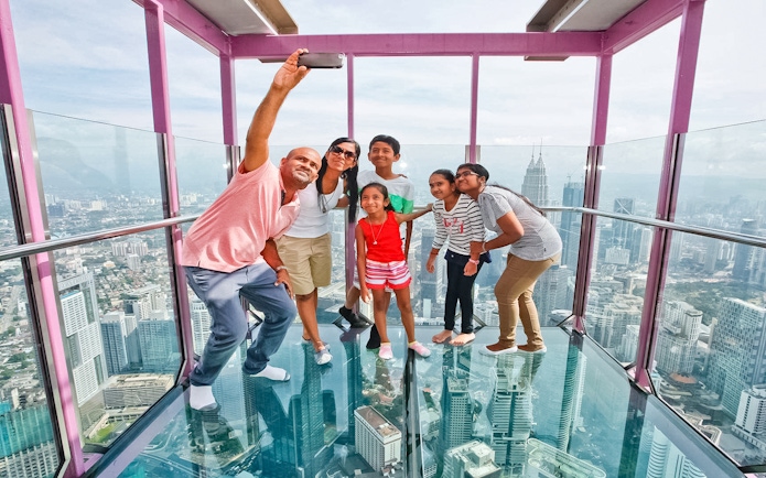 Family taking a selfie on the Sky Box at KL Tower, Malaysia, with cityscape views.