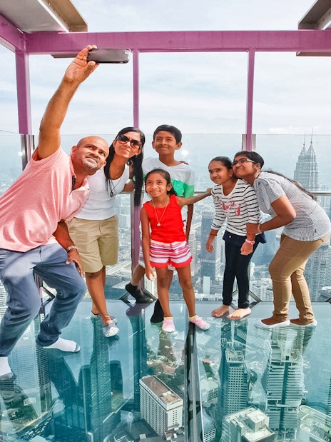 Family taking a selfie on the Sky Box at KL Tower, Malaysia, with cityscape views.