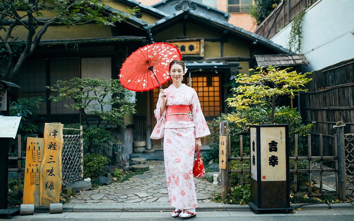 Kimono-clad woman with red umbrella in front of traditional Kyoto house.