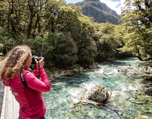 Person photographing a river on the Milford Track, surrounded by lush forest in Te Anau, New Zealand.