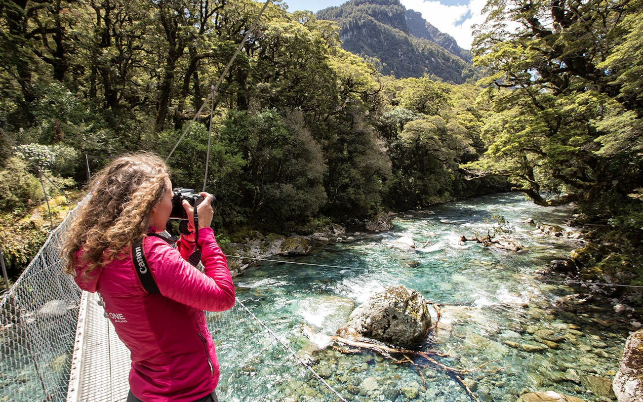 Person photographing a river on the Milford Track, surrounded by lush forest in Te Anau, New Zealand.