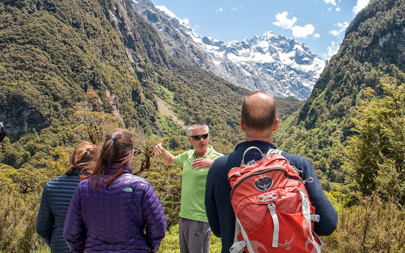 Group hiking with guide in green shirt at Lake Marian, New Zealand, surrounded by mountains.