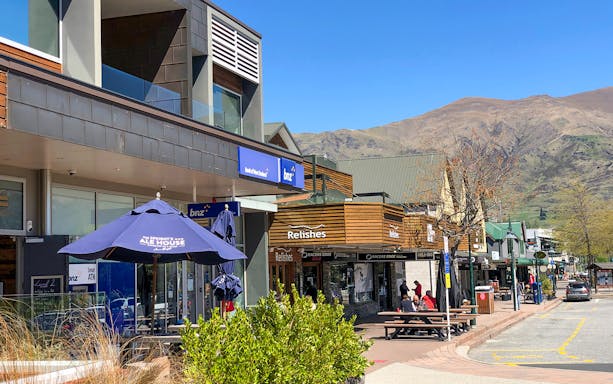 Shops and cafes on a sunny street in Arrowtown, New Zealand, with mountains in the background.