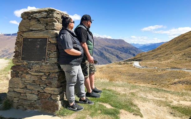 Two people standing by a stone monument overlooking a scenic mountain view on the Arrowtown and Wanaka Highlights Tour.