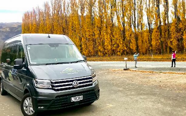 Tour van parked near autumn trees in Arrowtown, New Zealand, with tourists taking photos.