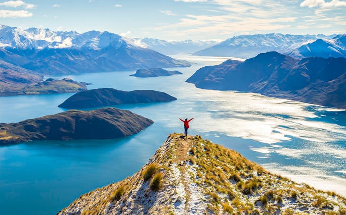 Person standing on a mountain peak overlooking Lake Wanaka and surrounding mountains, New Zealand.