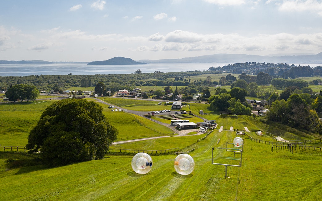 Zorb giant inflatable balls rolling down a grassy hill with a scenic view of Lake Rotorua, New Zealand.