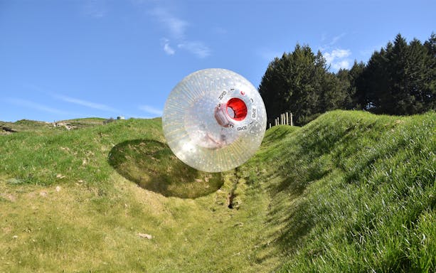 Zorb ball rolling down a grassy hill under a clear blue sky.