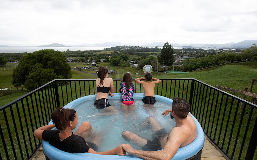 Family in hot tub overlooking landscape with Zorb ball ride in the distance, Rotorua, New Zealand.