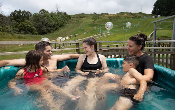 Family relaxing in a hot tub with Zorb inflatable balls on a hillside in the background.