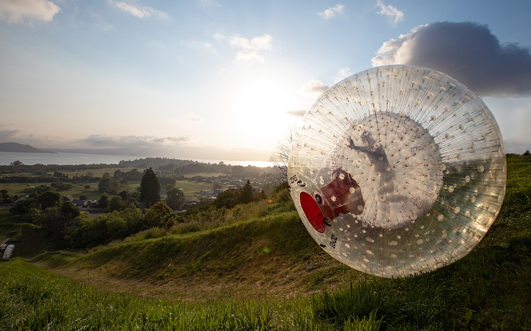 Person rolling downhill in a giant inflatable Zorb ball at sunset, overlooking a scenic landscape.