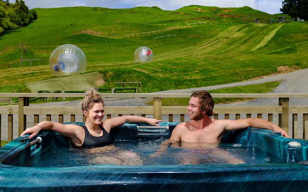 Couple relaxing in a hot tub with Zorb inflatable balls on a grassy hill in the background.