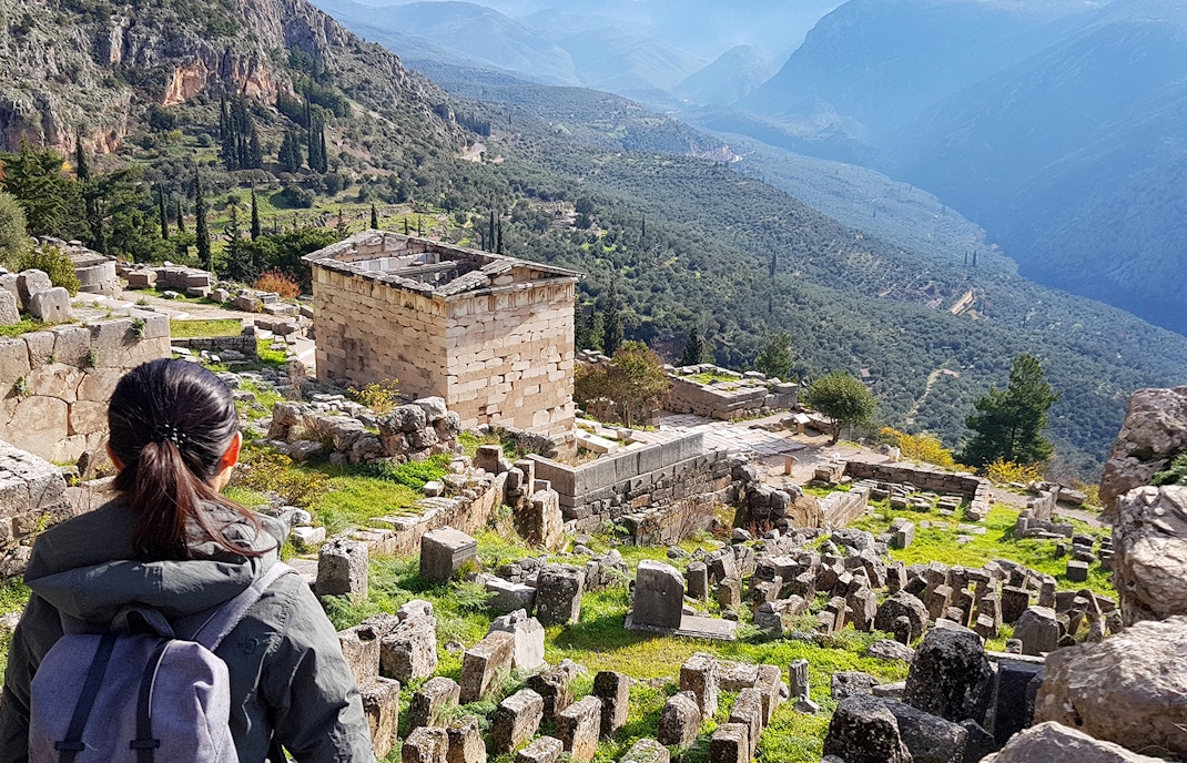 Visitor exploring ancient ruins at Delphi Archaeological Site, Greece, with mountainous landscape.