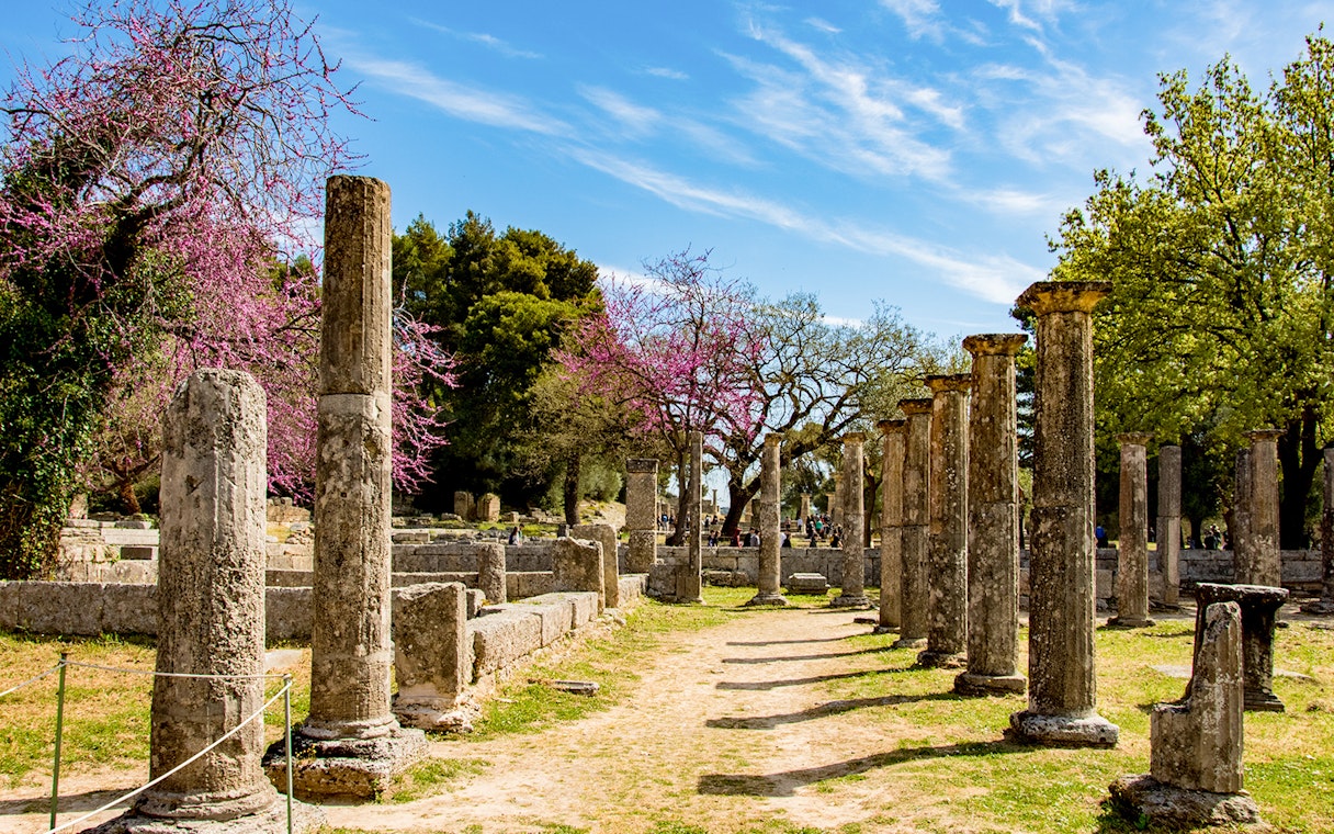 Ancient stone columns at Olympia archaeological site, Greece, with blooming trees in the background.