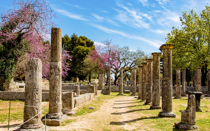 Ancient stone columns at Olympia archaeological site, Greece, with blooming trees in the background.