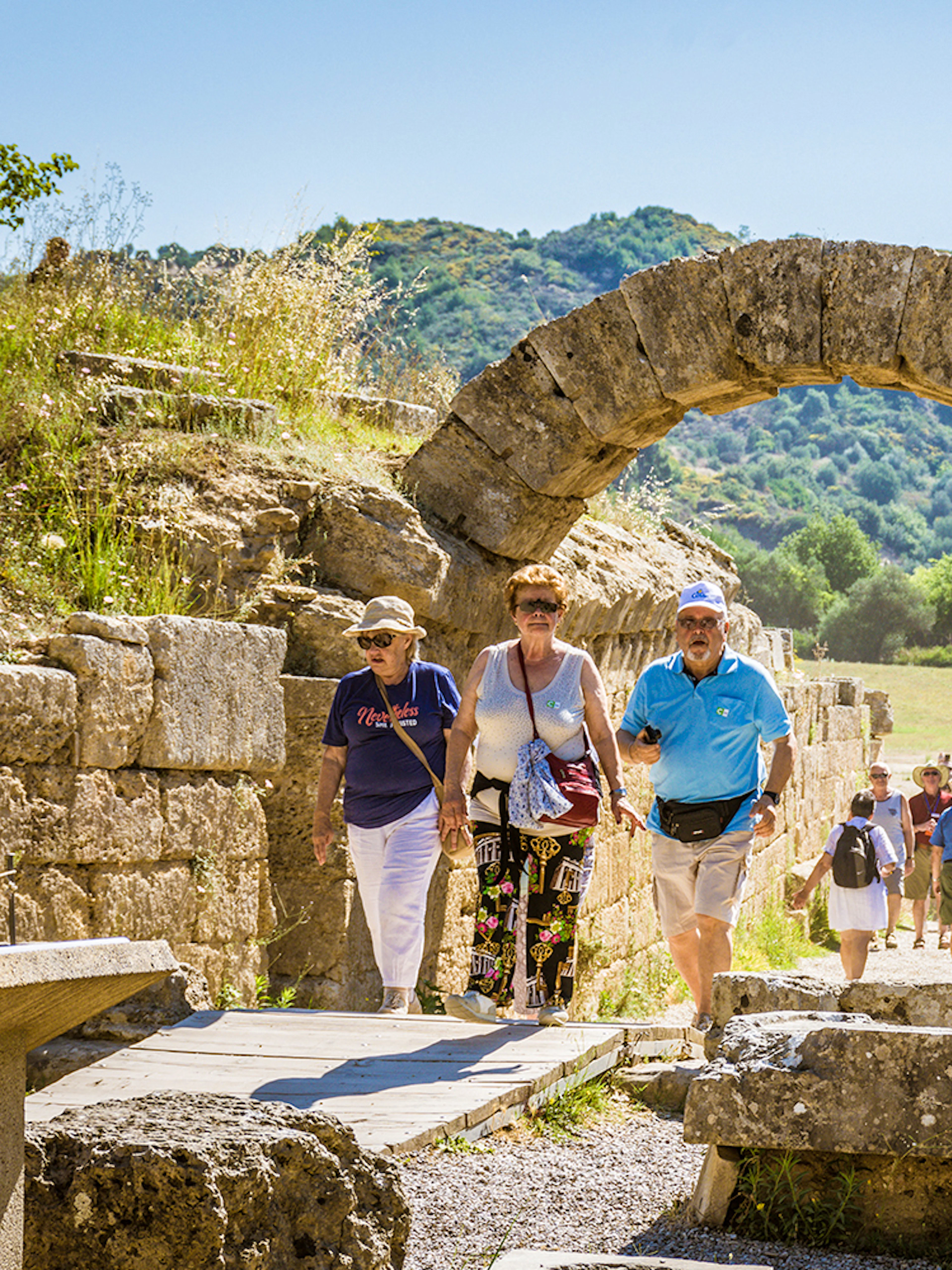 Visitors walking through ancient stone archway at Olympia archaeological site.
