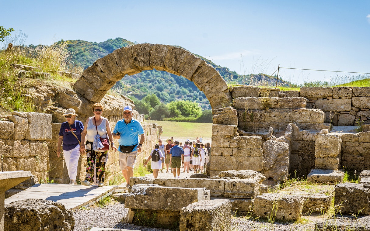 Visitors walking through ancient stone archway at Olympia archaeological site.