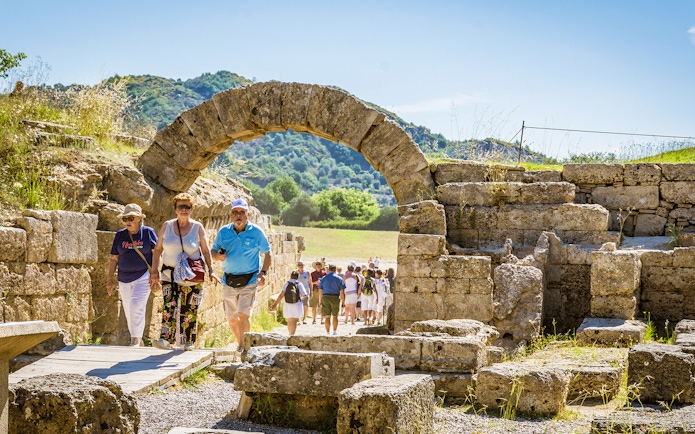 Visitors walking through ancient stone archway at Olympia archaeological site.