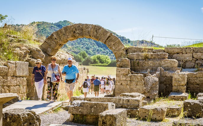 Visitors walking through ancient stone archway at Olympia archaeological site.