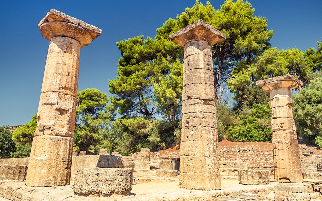 Ancient stone columns at Olympia archaeological site, Greece, surrounded by trees.