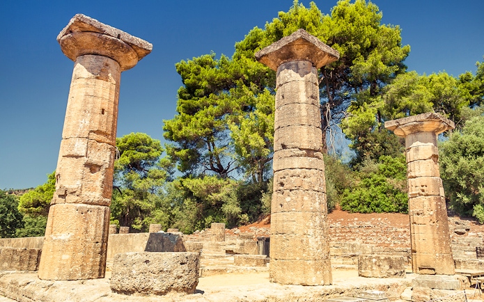 Ancient stone columns at Olympia archaeological site, Greece, surrounded by trees.