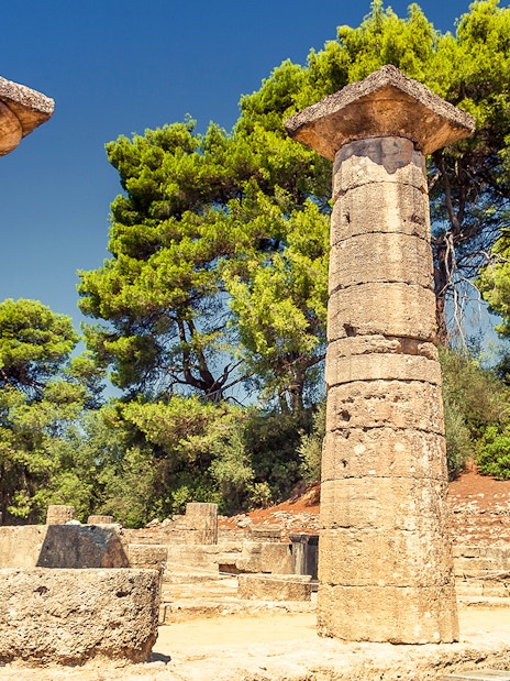Ancient stone columns at Olympia archaeological site, Greece, surrounded by trees.