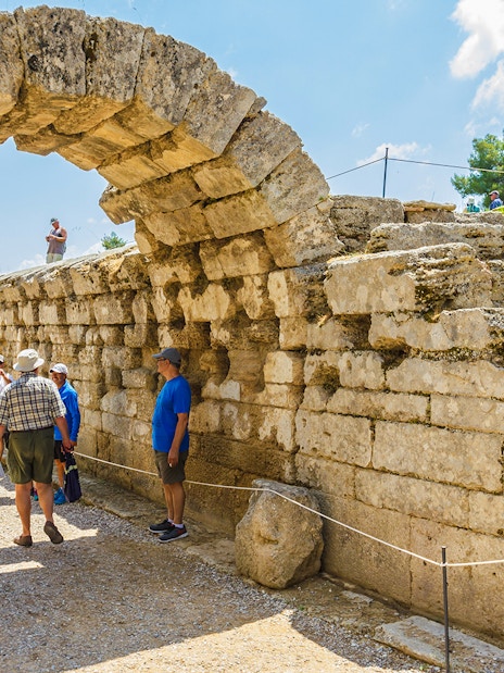 Visitors exploring the ancient stone archway at Olympia Archaeological Site, Greece.
