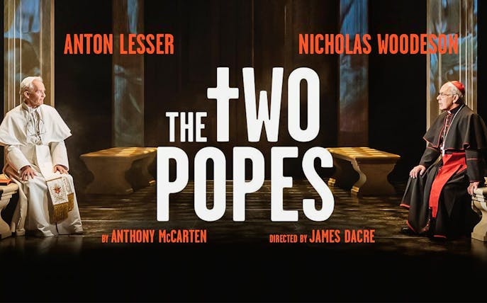 Two men in religious attire seated opposite each other in a dimly lit room, text reads "The Two Popes.