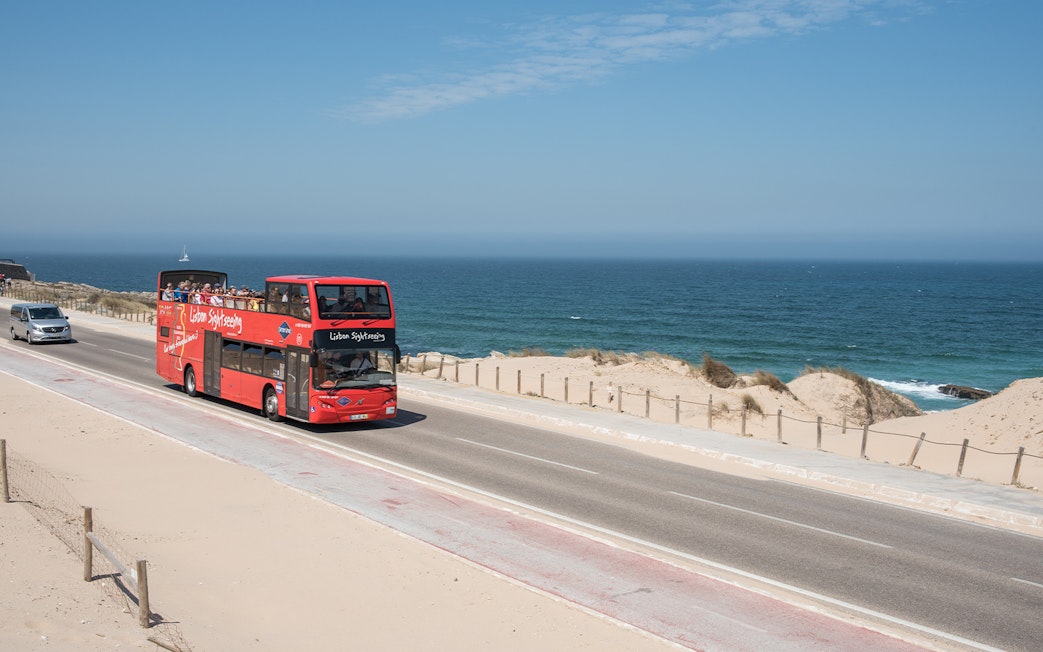 Red double-decker bus on Lisbon coastal road with ocean view.