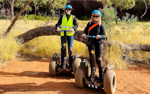 Tourists riding Segways on a dirt path near Uluru, surrounded by trees and grass.