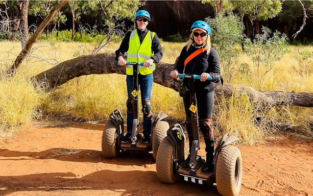 Tourists riding Segways on a dirt path near Uluru, surrounded by trees and grass.