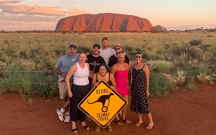 Group posing with Uluru Segway Tours sign in front of Uluru, Australia.