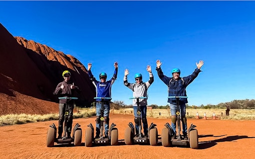 Tourists on Segways near Uluru, Australia, with arms raised against a clear blue sky.
