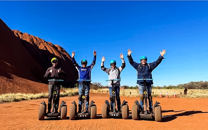 Tourists on Segways near Uluru, Australia, with arms raised against a clear blue sky.