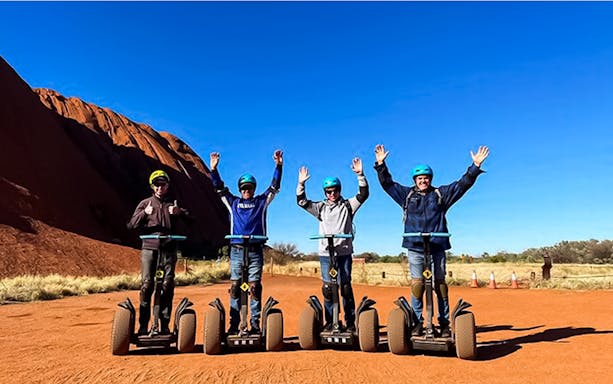Tourists on Segways near Uluru, Australia, with arms raised against a clear blue sky.