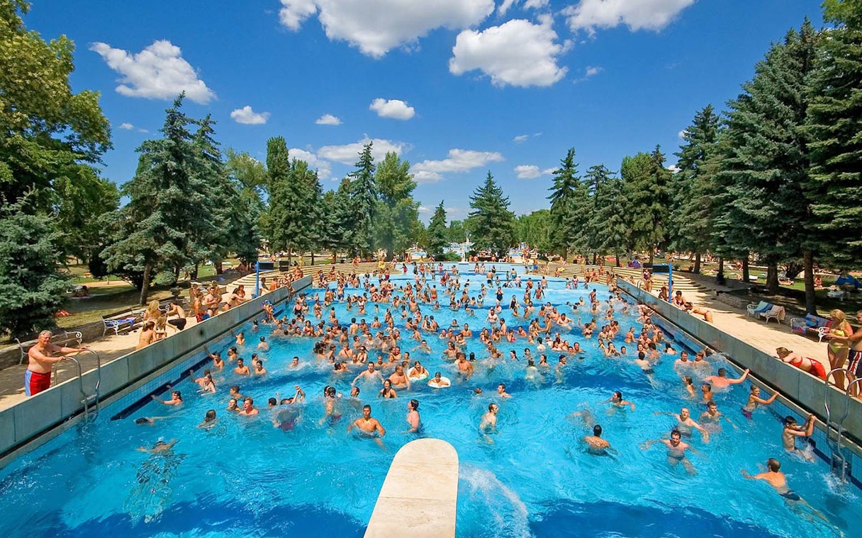 Crowded outdoor pool at Palatinus Spa, Budapest, surrounded by trees and sunbathers.