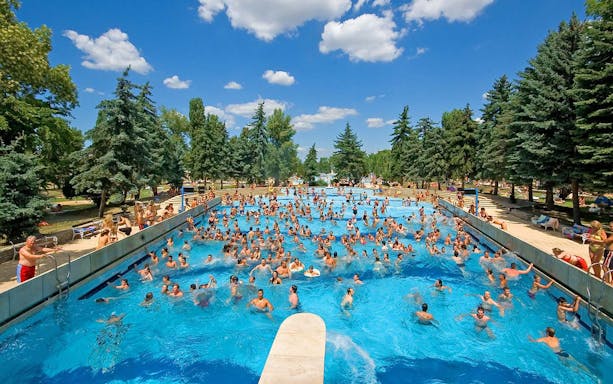 Crowded outdoor pool at Palatinus Spa, Budapest, surrounded by trees and sunbathers.