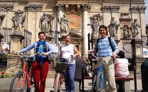 Three people with bicycles in front of a historic building with statues during a city bike tour.
