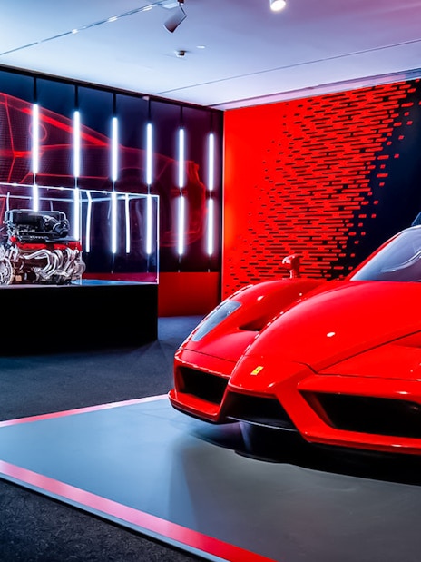 Red Ferrari Enzo displayed at Ferrari Museum Maranello with engine exhibit in background.