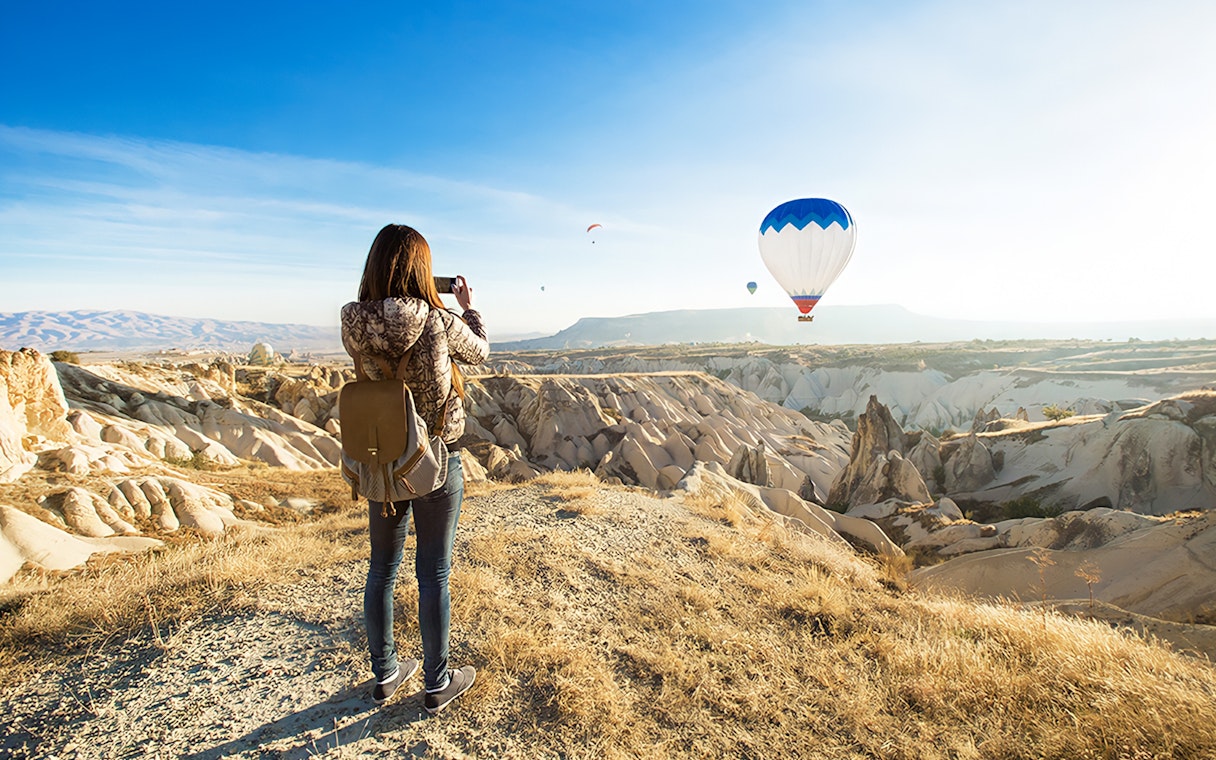 Person photographing hot air balloons over Cappadocia's unique rock formations.