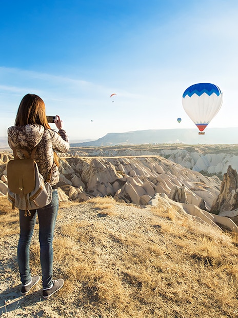 Person photographing hot air balloons over Cappadocia's unique rock formations.