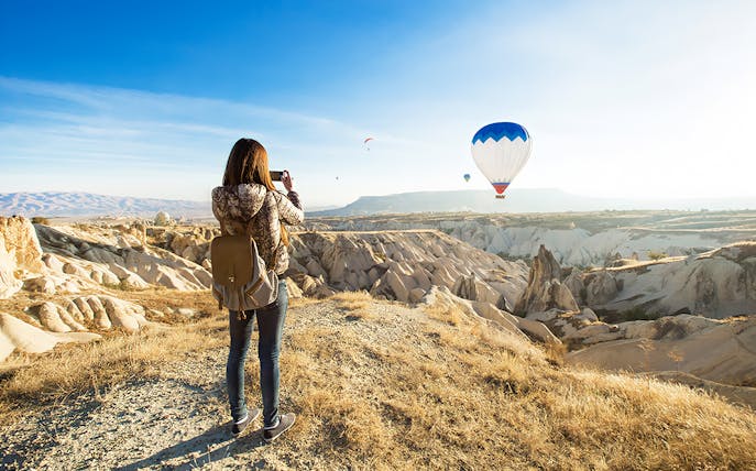 Person photographing hot air balloons over Cappadocia's unique rock formations.