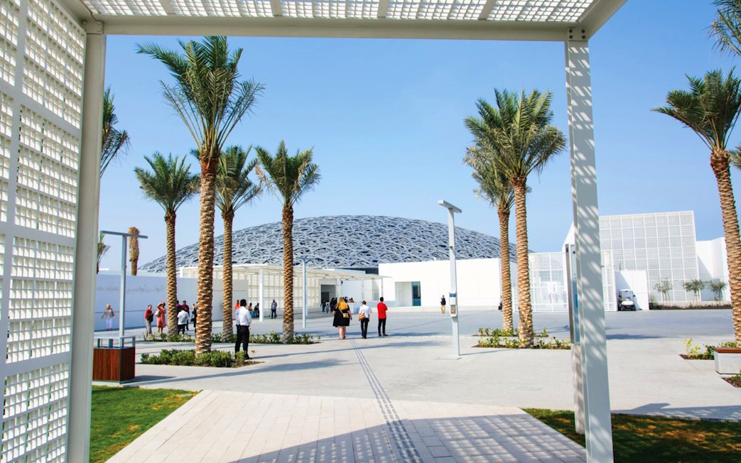 Louvre Abu Dhabi exterior with visitors walking under palm trees.