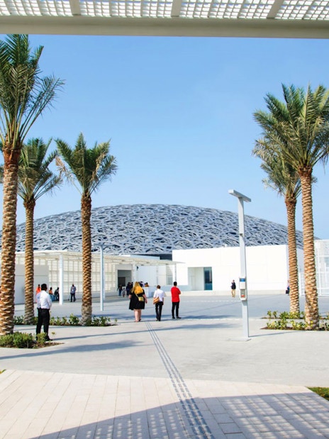 Louvre Abu Dhabi exterior with visitors walking under palm trees.