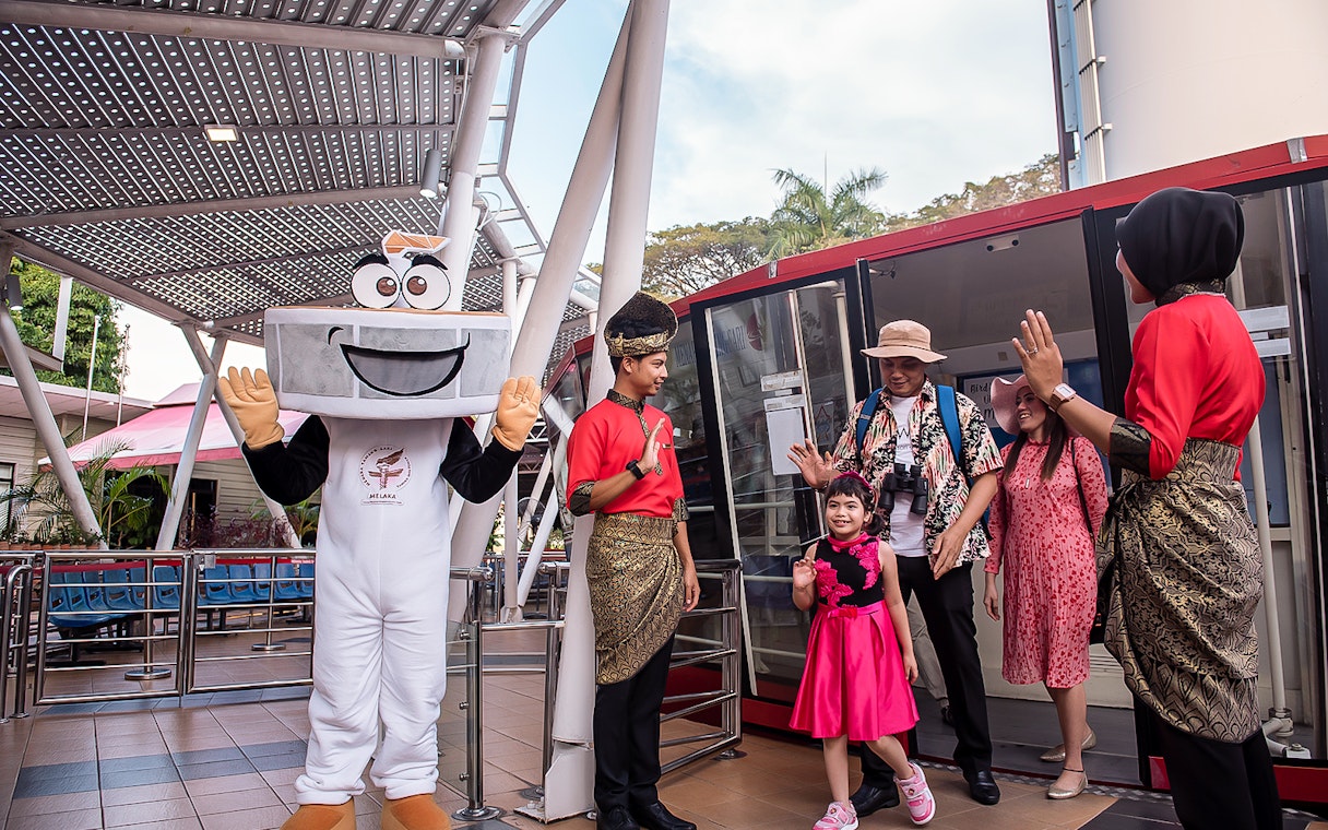 Visitors at Menara Taming Sari, Melaka, greeted by a mascot and staff in traditional attire.