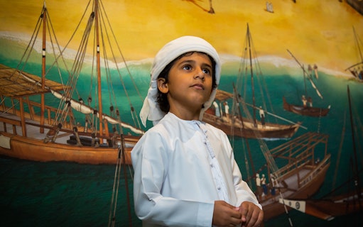 Child in traditional attire at Al Shindagha Museum with dhow mural in background.