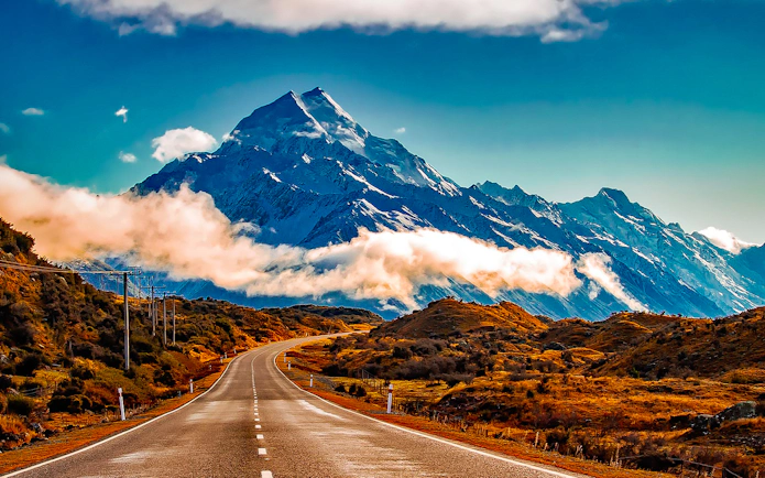 Road leading to Mt Cook with snow-capped peaks and clouds, New Zealand.