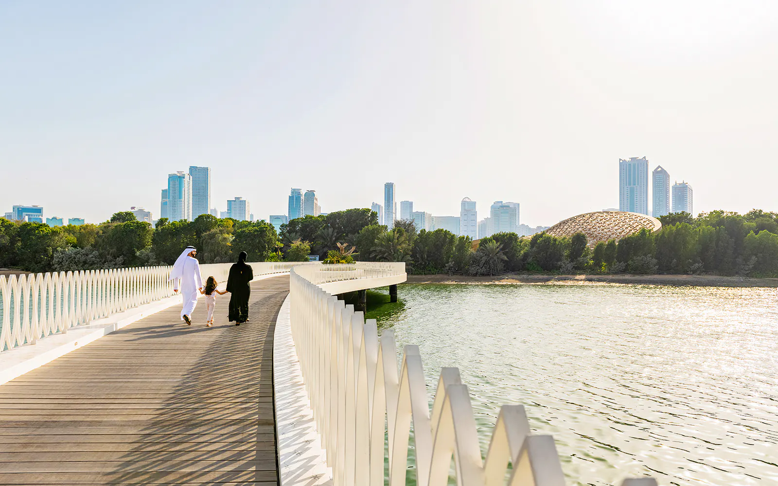 A family walks along the white bridge to Al Noor Island with a domed pavilion across the water.