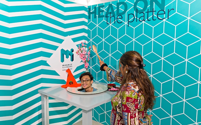 Person interacting with head-on-a-platter illusion at Museum of Illusions Kuala Lumpur.