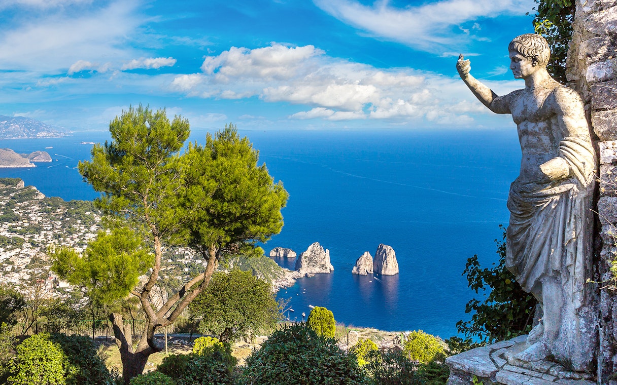 Capri coastline view with Faraglioni rocks and statue, seen from a hillside vantage point.